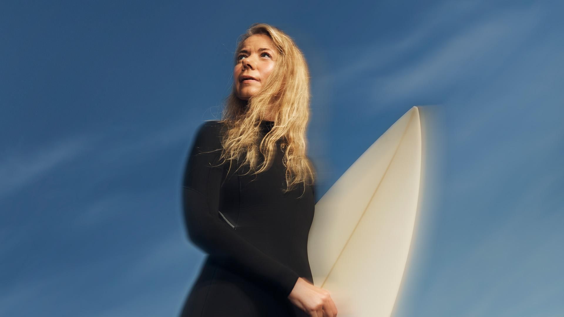 A woman wearing a black wetsuit holds a surfboard under blue skies, looking hopeful and determined. 