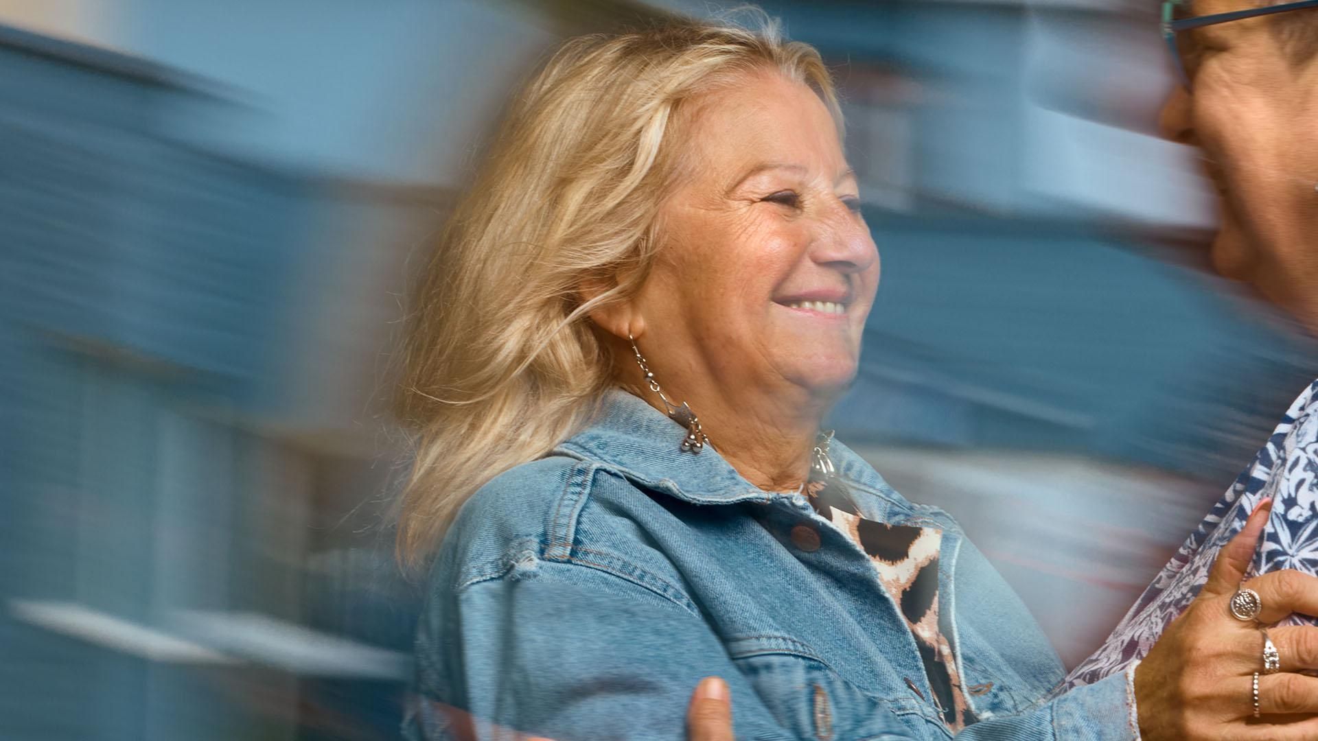 A smiling woman in a blue denim jacket reaches out to embrace another woman wearing glasses. Overlay text reads: "Expert patient and mentor."​