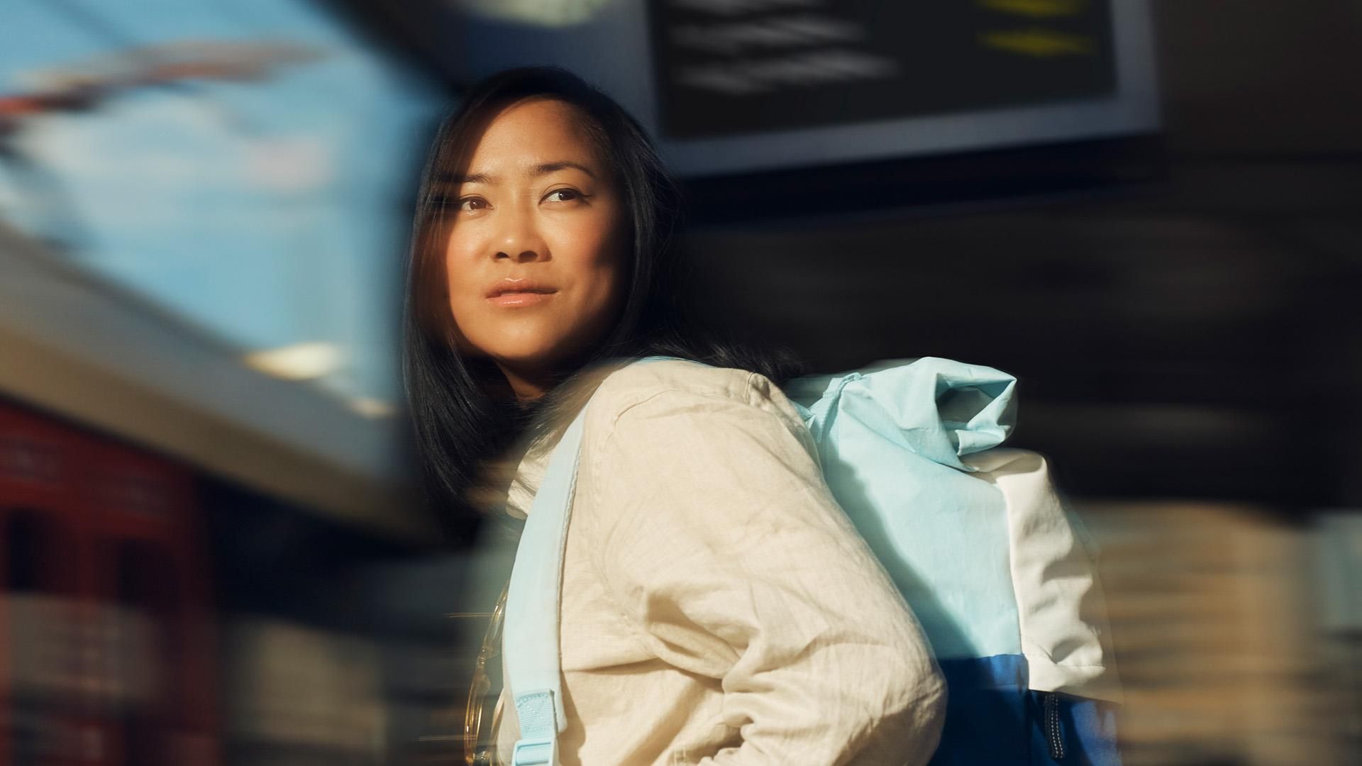 A woman wearing a yellow shirt and blue backpack gazes into the distance with a dreamy expression. Overlay text reads: "Adventurer."​