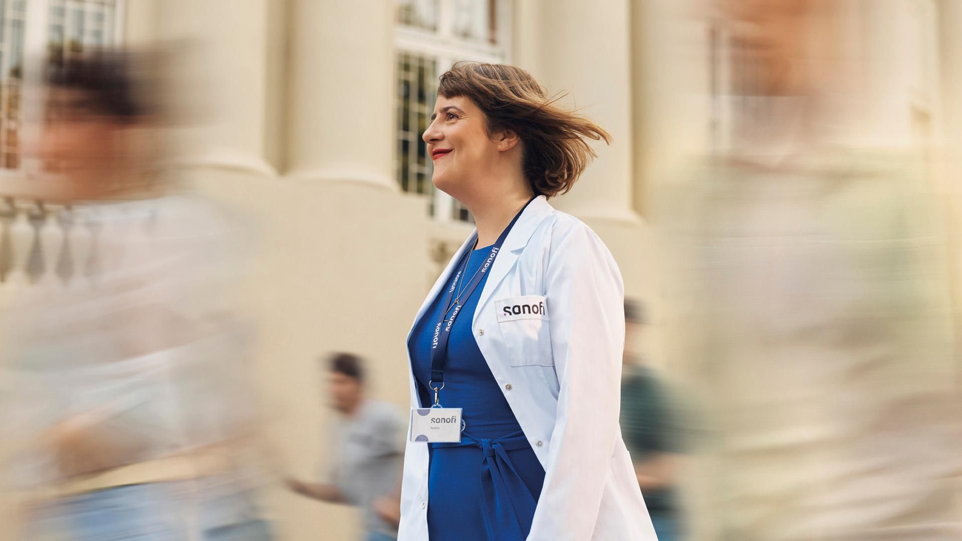 A smiling woman wearing a white Sanofi lab coat and company badge smiles while walking forward outside of a building. Several people are running alongside her in the background, they are blurred in motion. Overlay text reads: "Working in Research and Development."​